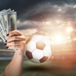 close-up-man-s-hand-holding-us-dollars-against-background-stadium-concept-sports-betting-making-profit-gambling-208405993